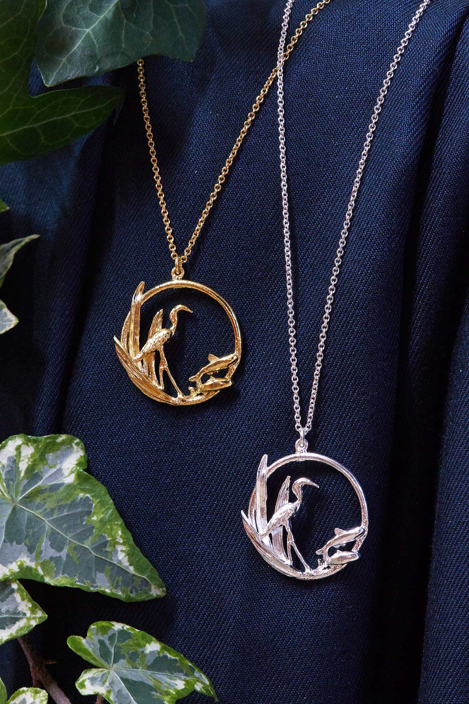 Two Heron & Fish Loop silver and gold plated necklaces hang against blue fabric with ivy