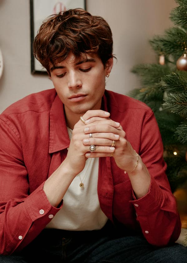 Man wearing a red shirt with a selection of rings including a spinnin signet ring. He wears a long chain disk necklace and single small hoop earring.