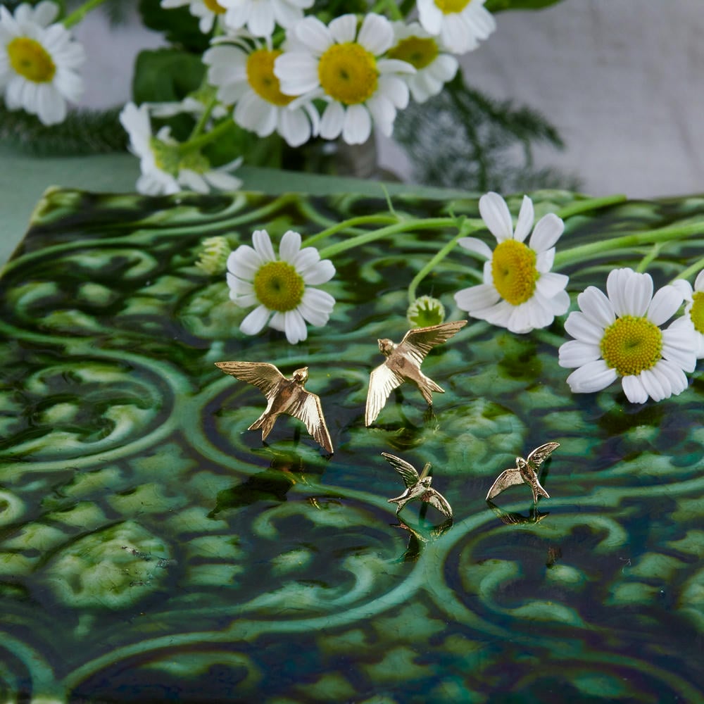Hummingbird and Swallow gold earrings on a green printed tile, surrounded by daisies