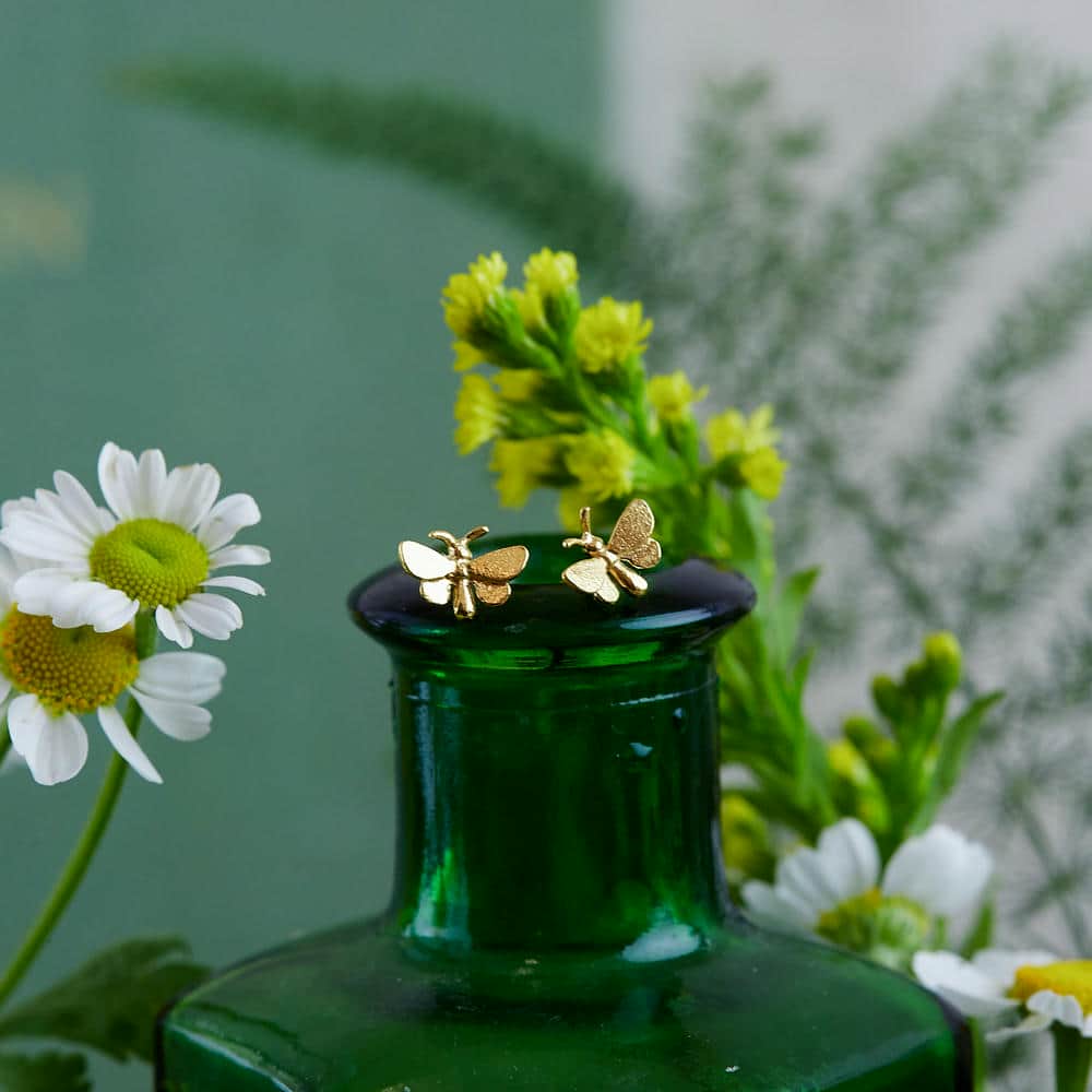 Pair of tiny gold butterfly stud earring perched atop a dark green glass bottle with daisies and yellow flowers surrounding.