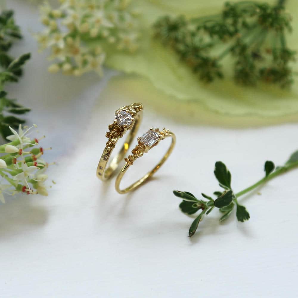 Two 18ct solid yellow gold engagement rings, one with a emerald cut diamond, the other a round brilliant cut, both with floral details, surrounded by pale white flowers and green foliage all on white parchment paper.