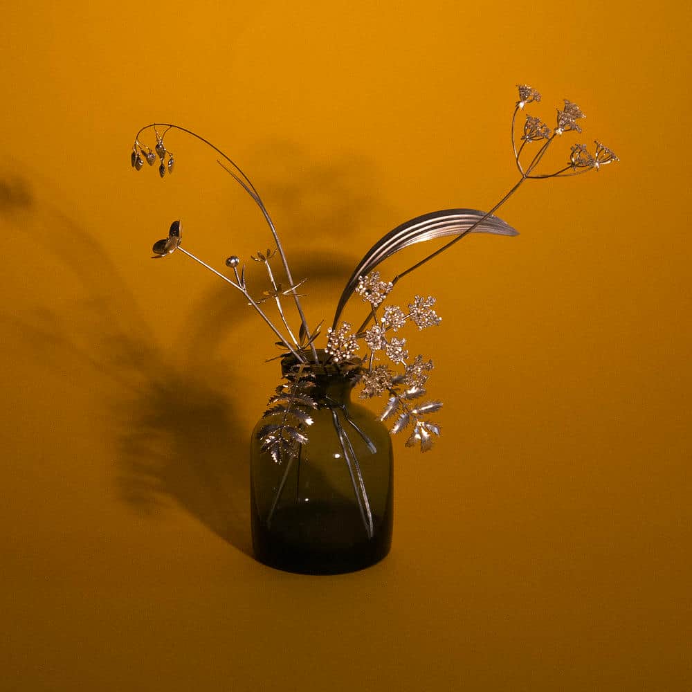 Sculptural silver flower and leaf stems arranged in a smoke coloured glass bottle against an orange backdrop.