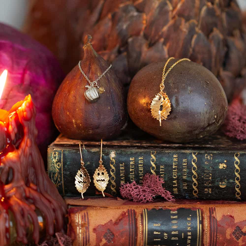 Silver pumpkin necklace, gold spooky leaf necklace and earrings displayed on old dark coloured books with a dark red melting candle.