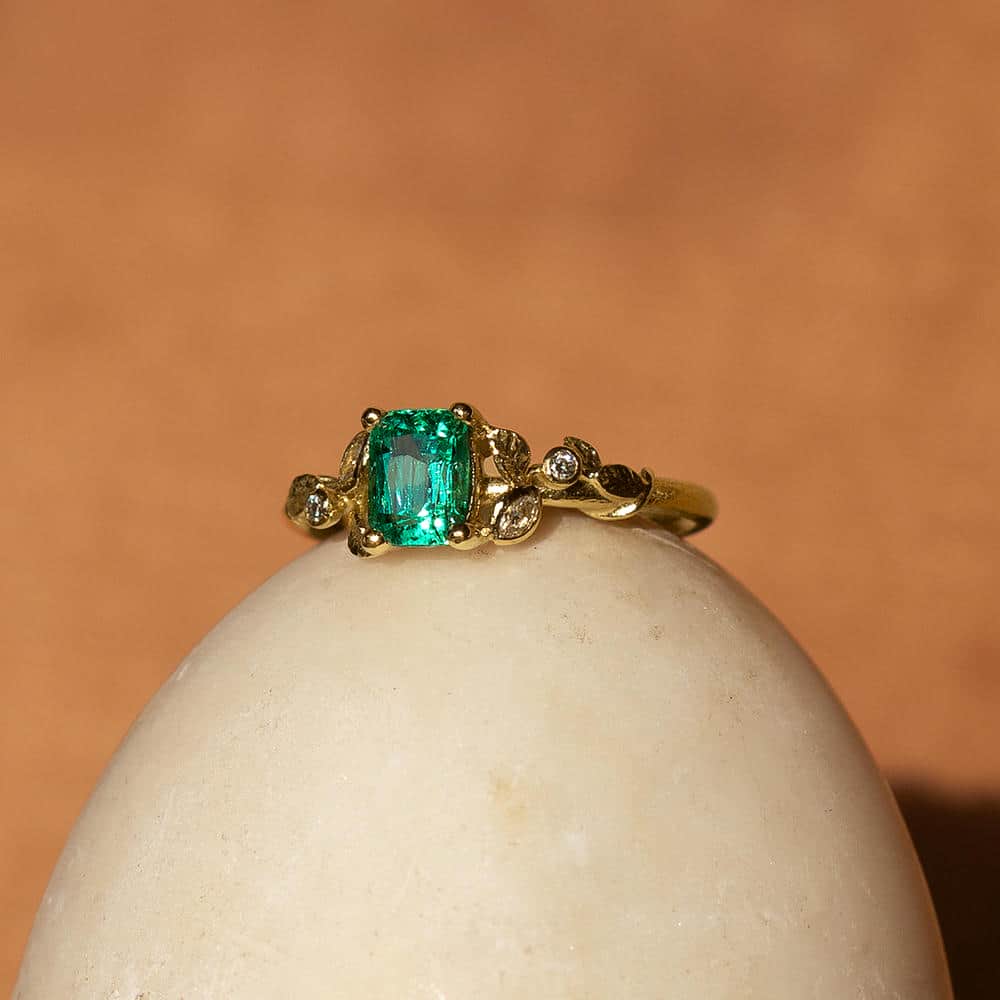 Solid yellow gold ring with leaf and small diamond design surrounding a large rectangular bright green emerald gemstone. The ring is displayed on top of a marble egg, against a mottled terracota orange backdrop.
