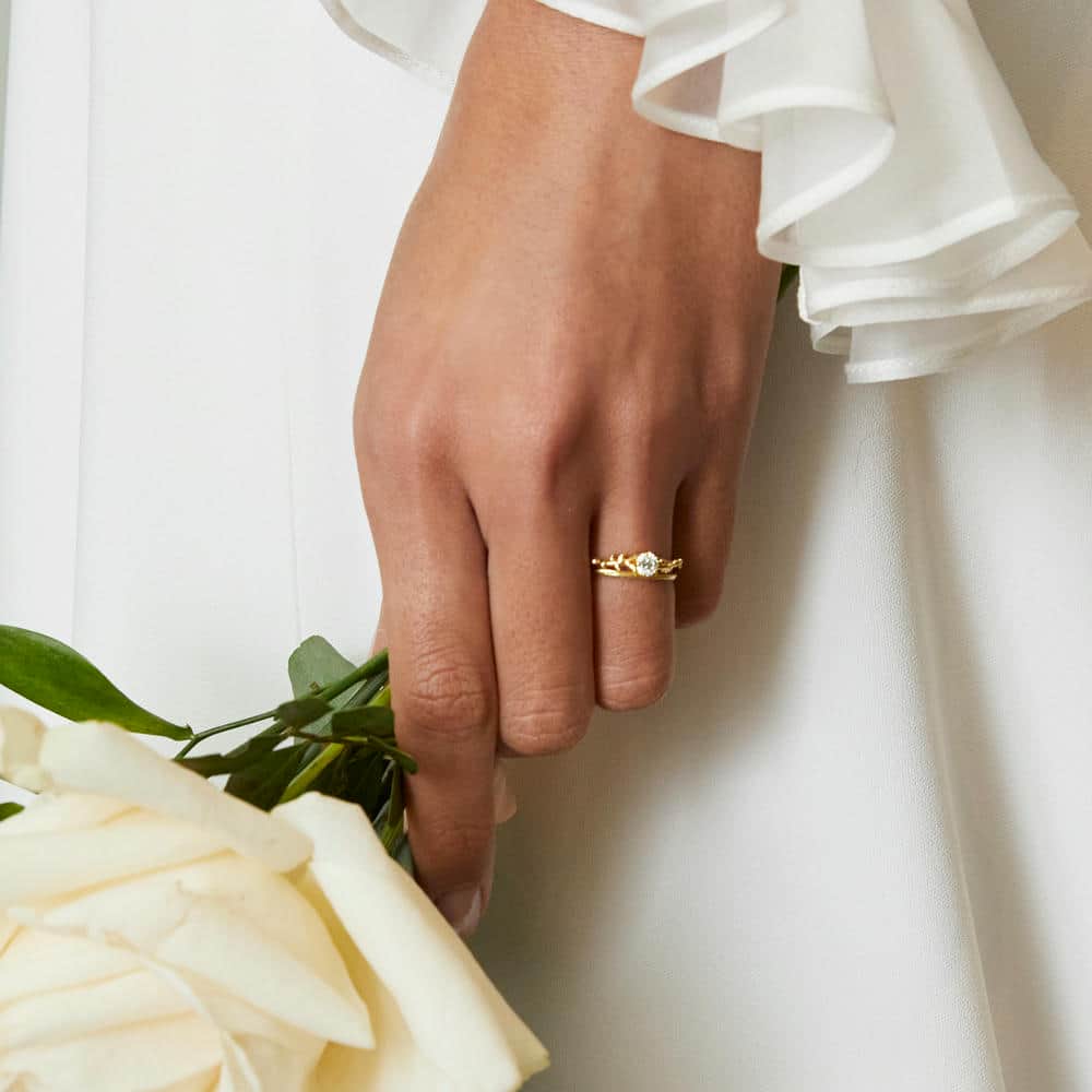 Woman wearing white flowing dress with wavy semi-transparent sleeve, holding a white rose and wearing solid 18ct yellow gold engagement and wedding rings.