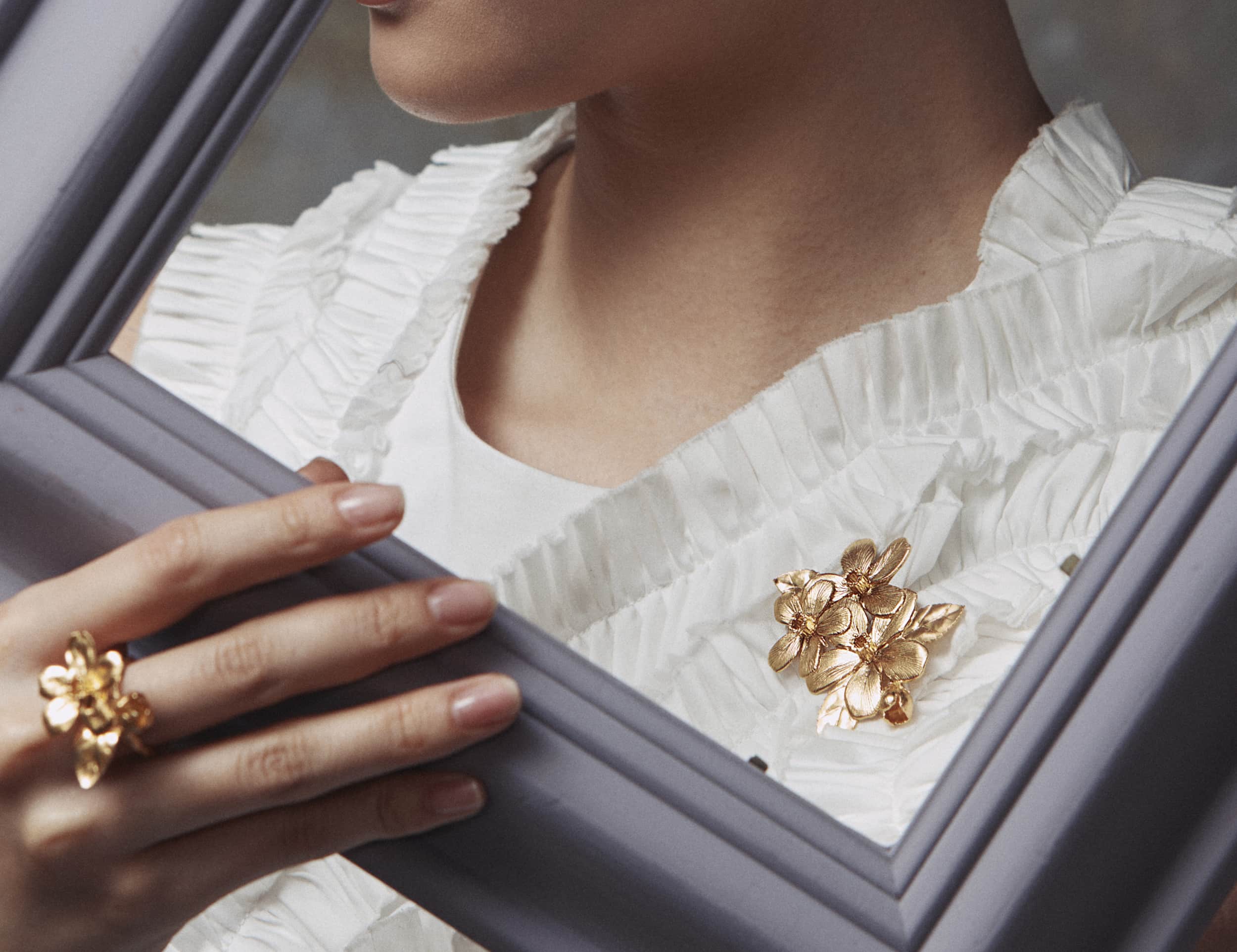Woman holding the outside of a lilac frame at an angle in front of her self to frame her neck, white ruffled dress and large floral gold brooch.