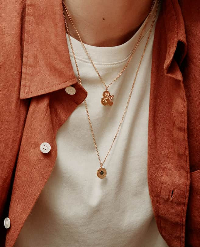 Model wearing a coral and white top, wears a Alex Monroe's ampersand necklace layered with a Raven Smith Iris Reversible Disc Green Tourmaline Necklace