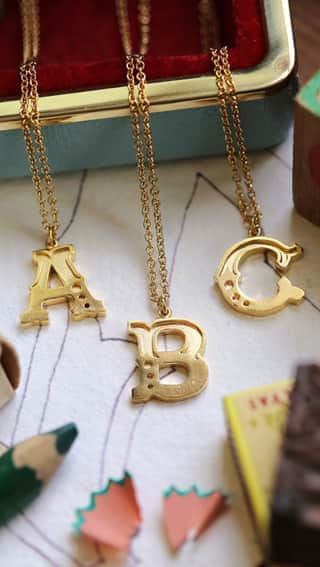 Just My Type Collection, Initial Necklaces A, B and C displayed on a writers desk with sketches and traditional writers implements including a pencil, pencil sharpener and vintage metal storage tins.