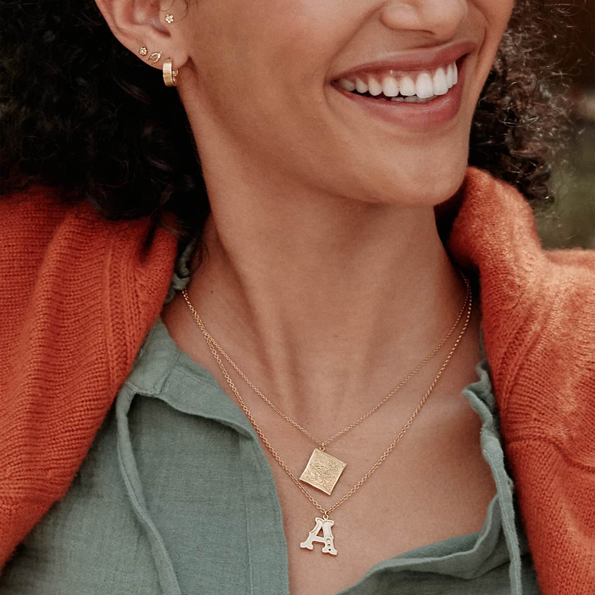 Model wearing a teal green top and a bright orange sweater is wearing Alex Monroe's gold plated letter, A Necklace and a Seed packet necklace.