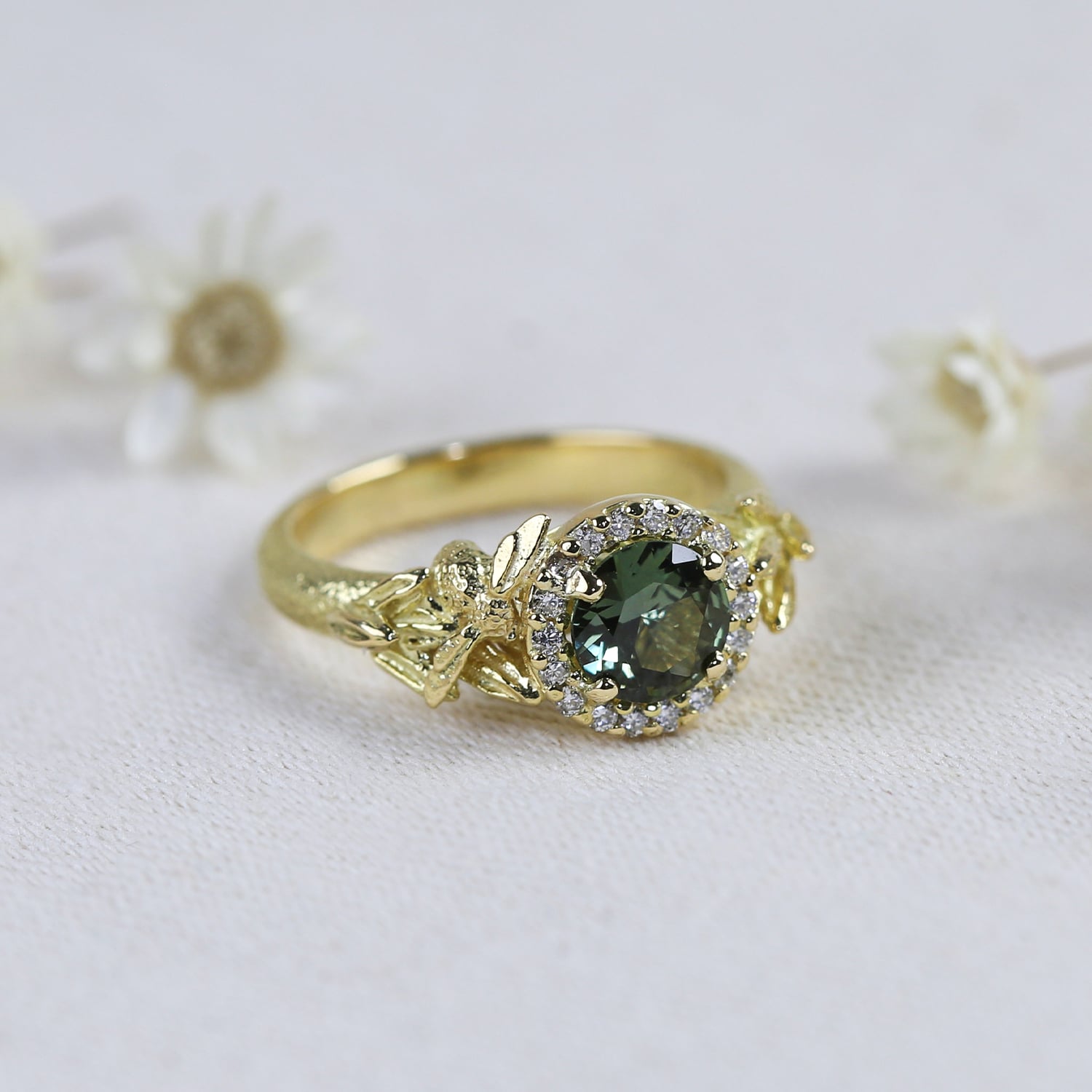 emerald gemstone 18ct yellow gold bespoke design engagement ring shown held in jewellers pliers against white petals