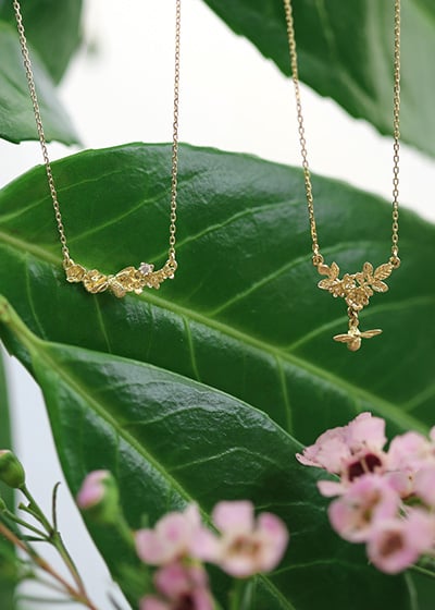 Floral Cluster Necklace with Bee Drop and Floral Curve Diamond Necklace hanging on top of a large green leaf