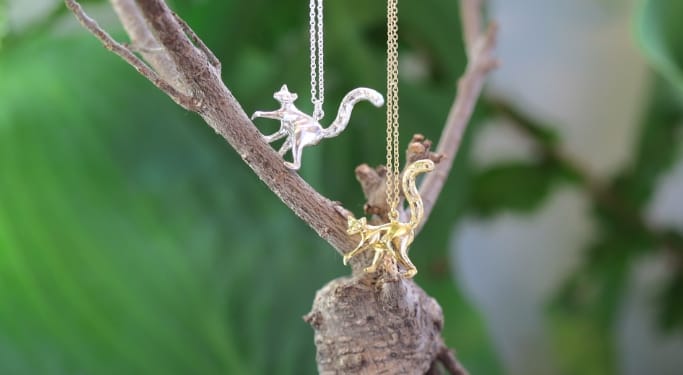 A silver and a gold plated Lemur Necklace both climbing a green plant.