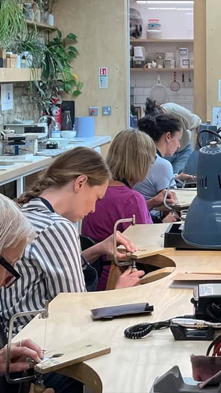 A Jewellery School class in action at the Alex Monroe workshop with attendees sat at jewellery benches using handsaws to cut out silver shapes.