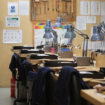 Tidy jewellery workbenches each with a blue anglepoise lamp and a tray containing drawing supplies and tools for Jewellery School attendees.