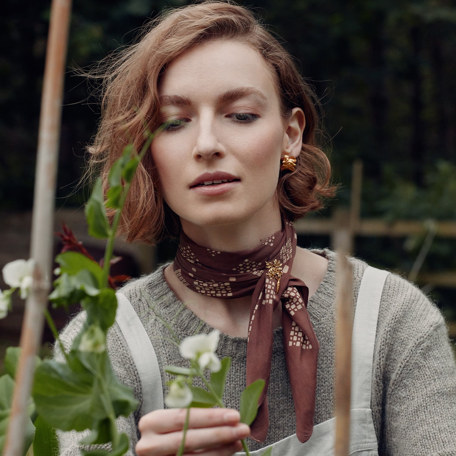 Model wearing Alex Monroe's Gold plated Chard Leafy Hoop Earrings and a gold plated Leafy Carrot Pin Brooch pinned into a brown scarf